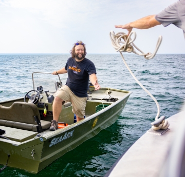 A person on a small boat prepares to grab a line being tossed by someone on another boat. His shirt says "I Am Buffalo State"