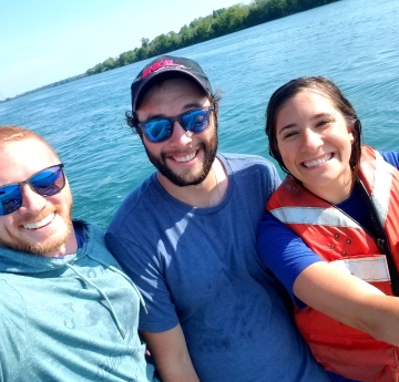 Three smiling people stand in front of the water. Two men wearing sunglasses and a woman in a life jacket