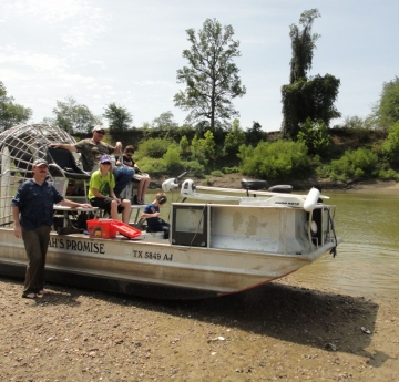 A boat with a large fan on the back sits on the sand. There are a few people on the boat, including children. One person stands on land next to the boat, and one person stands in the water.