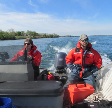 Two people sit at the back of a boat while one drives. There is a net next to one of them, and both are wearing safety suits.