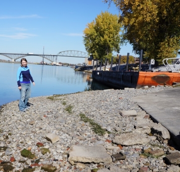 A person stands on the gravel by the waterline. The dock and concrete pad are about ten feet up the ramp. There is a line of weeds washed up about halfway between the water and the concrete pad, marking a previous waterline.