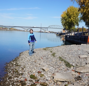 A person stands on the gravel by the waterline. The dock and concrete pad are about ten feet up the ramp. There is a line of weeds washed up about halfway between the water and the concrete pad, marking a previous waterline.