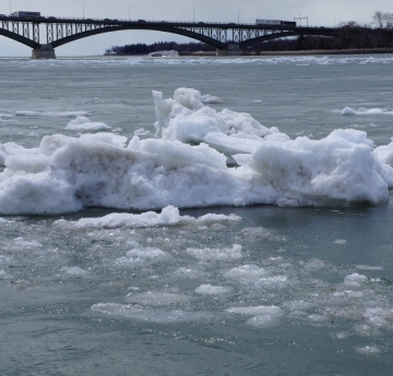 A large chunk of ice in an icy river. There is a bridge in the distance.