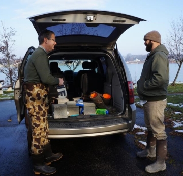 Two people stand at the open gate of a minivan with cinderblocks inside, in a parking lot by a river. There is some snow on the grass.