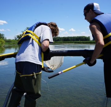 Two people stand at the front of a boat holding nets on long poles. One is looking at something in their net, while the second glances over at the net too.