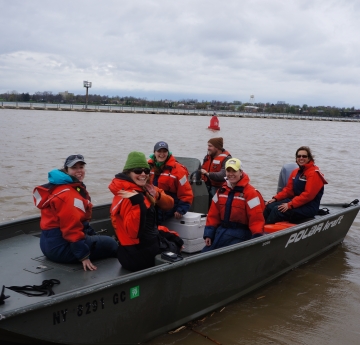 Six people in coldwater safety gear on a small boat in a muddy river on a cold day. Most are smiling or waving at the camera while one drives