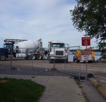 A fence with a sign "DANGER KEEP OUT" blocks a construction zone with two cement trucks and some other equipment. There is a body of water behind the construction zone.