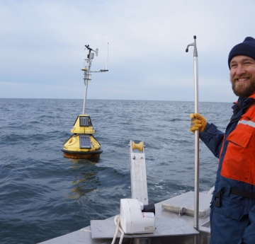 A smiling person in winter gear stands on the back deck of a boat, holding a boat hook. There is a buoy in the water nearby and a pot puller winch on the side of the boat.