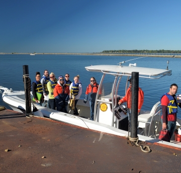 A group of people stand on a fiberglass boat tied up to a dock. Two people on the dock hold the lines.