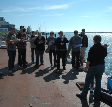 A group of people stand on a dock by the water taking notes. A person stands in front of them with some research equipment