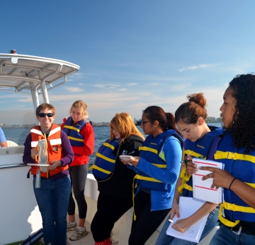 Several people wearing life jackets take notes on a boat while a person holds up a disk with a rope attached to it. Another person sits at the helm of the boat.