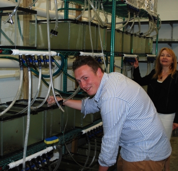 A person smiles and stands by a shelf with many aquariums full of water in a lab while a second person also smiles and stands nearby
