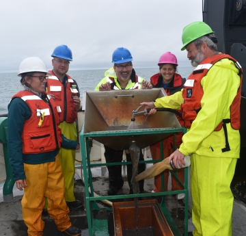 Five people stand around a metal basin on the back of a boat. One is spraying a hose into the basin.