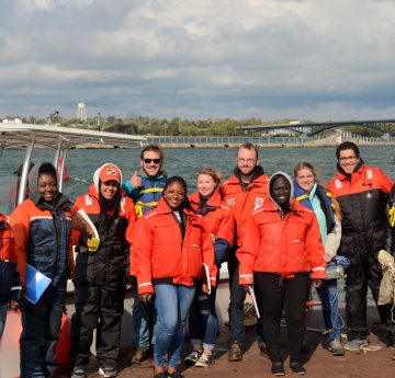 A group of ten people wearing cold water safety gear standing in front of a docked boat by the water.