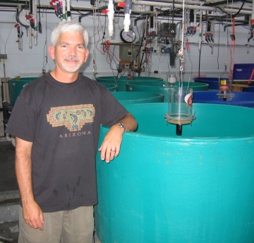 A person stands with their arm resting on a large round tank in a wet lab. Their shirt says "Arizona."