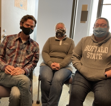 Three smiling people wearing face masks sit in chairs next to each other in a small office. There is an open door behind them.