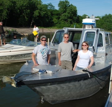 Three people stand for a picture at the front of a metal boat that's tied up to a dock. There are some other people launching a boat at the next slip over.