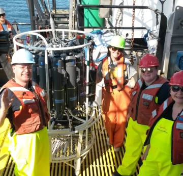 Four people stand on the deck of a large boat next to an instrument made of cylindrical bottles in a frame