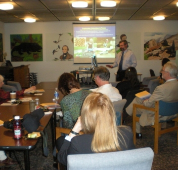 a group of people in chairs watch a presentation given by a person at the front of the room. There is a presentation projected onto a screen, and murals painted on the walls of the room.