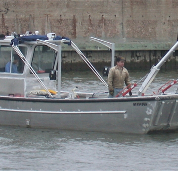 One person works in the front of a boat with an A-frame on the front of it. Another person is in the cabin.