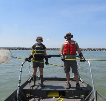 Two people stand in the front of a boat wearing life jackets, ear protection, rubber gloves and rubber boots, and holding nets on long poles.