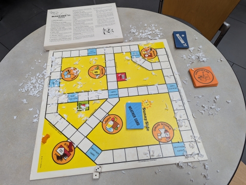 A vintage board game, "Blizzard of '77," set up on a table with some fake snow. The board has intersecting roads with errand destinations and different colored cards. There are destination cards like "Hardware store," "Blizzard '77" cards, car tokens, and two dice, along with the rules on the box insert.