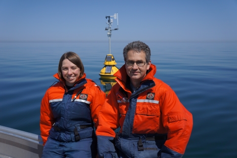 Two people in safety suits sit on the edge of a boat on a flat calm day with a observation buoy behind them