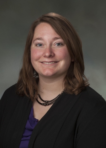 A person with shoulder-length brown hair and a ruddy complexion smiles, wearing a black cardigan over a purple blouse, and a necklace