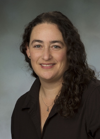 A person with dark, long, curly hair and light-toned skin smiles, wearing a dark blouse and a necklace with a pendant