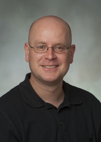 A bald person with light-toned skinned and wire-framed glasses with small oval lenses smiles, wearing a black polo