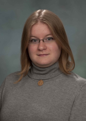A person with blond shoulder-length hair and glasses, wearing a light gray turtleneck and a short necklace with a round copper pendant with a compass rose.