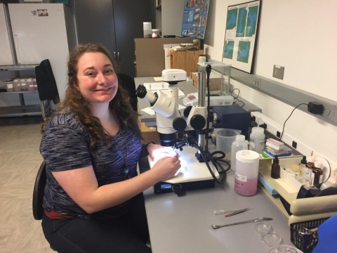 A person with long curly hair smiles and sits at a microscope in a lab.