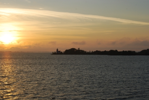 Sun refracting through wispy clouds in the morning above a spit of land with a lighthouse in a large lake.