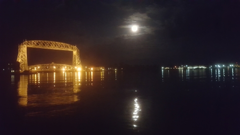 A night-time picture with a lighted steel liftbridge at left, a full moon at right, and the reflection of both in the water.
