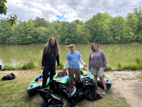 Three people smile and stand by kayaks and three garbage bags in front of a small lake