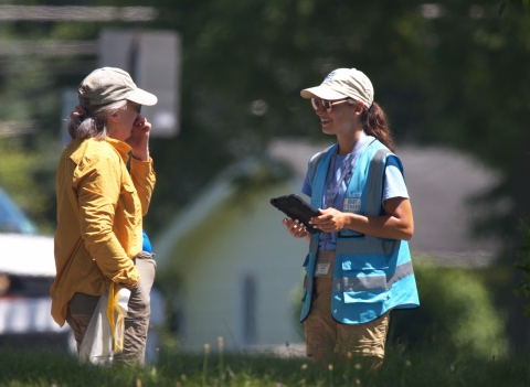 A person in a vest that says "boat steward" approaches a person outside to ask survey questions on a tablet.