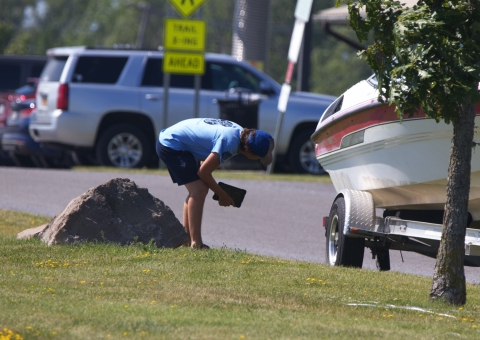 A person with a tablet crouches to inspect a boat and trailer in a parking lot.
