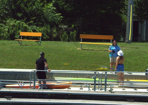 A person in a blue uniform vest with a tablet talks with two kayakers at a kayak launch.