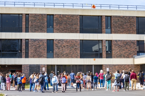 A crowd stands next to a three-storey classroom building as a person on a roof throws a pumpkin off a building, the painted pumpkin now about one storey above the ground.