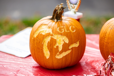 A decorated pumpkin sits on a table outside. The surface is scraped, not carved, in the shape of the Great Lakes with the letters G L C. Behind the pumpkin is a trophy.