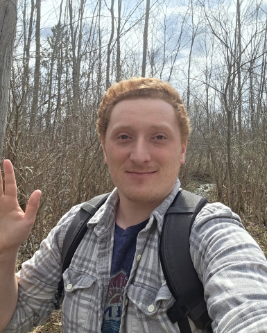 Richard Campbell waves and takes a selfie while hiking in a forest on a spring day with leafless trees