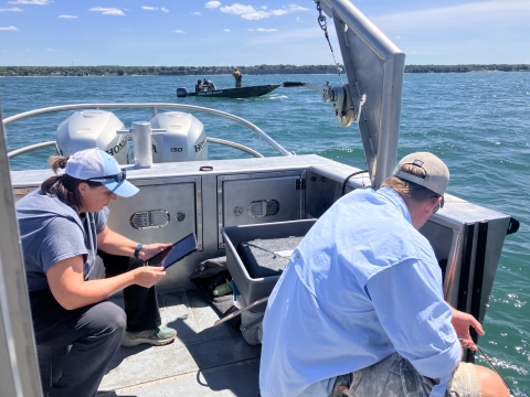 Two people work on the deck of a boat in a lake while three people work on a small boat in the distance.