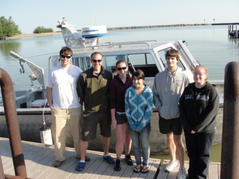 Six students pose on a dock next to a boat with a cabin.