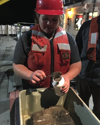 A person in a hard hat and life jacket stands in front of a tub with a square of mud, using a spoon to scoop some mud into a plastic jar.