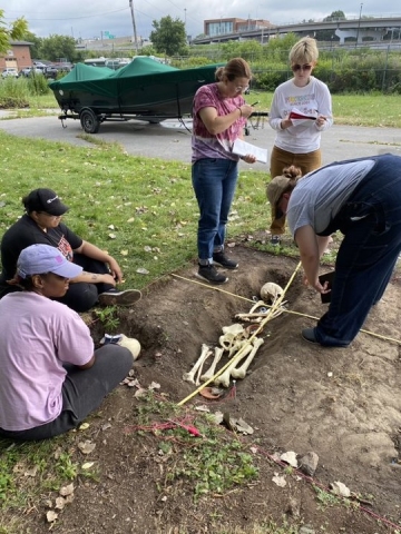 A few people sit or stand taking notes around a shallow hole in the ground with artificial human bones, marked with two measuring poles.