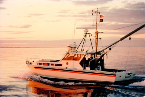 A boat with a cabin and an open back deck on a lake at sunset. A long boom arm extends from the back