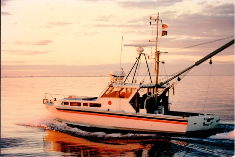 A boat with a cabin, lower deck, and long boom off the cabin sails through calm water during sunrise