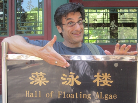 A person with short styled hair and glasses smiles and gestures to a sign they stand behind, which is labeled in pictograms and English as "Hall of Floating ALgae", with a building with fancy wood inlayed windows behind them