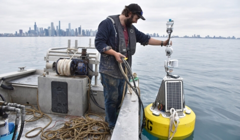 A person stands at the front of a boat holding onto the mast and a cable of a buoy. The Chicago skyline is in the background.