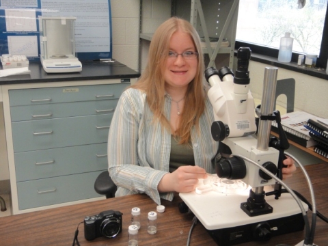 A person with long hair and glasses sits at a dissecting microscope in a lab with many vials nearby